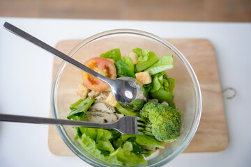 Chicken caesar salad with tomatoes, lettuce, broccoli and croutons in a glass bowl with fork and spoon, shot from above