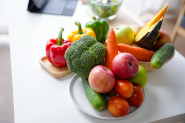 Fresh vegetables and fruits lying on a white table with a laptop in the background suggesting a healthy meal preparation