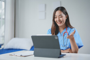 Smiling young female doctor is making a video call on a digital tablet, gesturing with her hand while sitting at a desk in her office