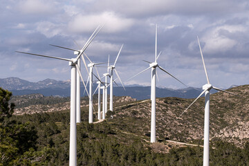 A line of wind turbines extends across a rugged hillside terrain, highlighting the use of renewable energy sources in a natural and challenging landscape in Tarragona Spain