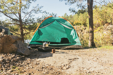 A tent on top of a mountain. Next to the tent a metal kettle is warming on a gas stove. Vacation in nature near the lake and pine trees.