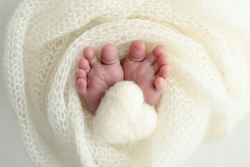 The tiny foot of a newborn baby. Soft feet of a new born in a white wool blanket. Close up of toes, heels and feet of a newborn. Knitted white heart in the legs of a baby. Macro photography. 