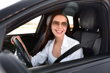 Smiling young woman in sunglasses with seatbelt driving car, view from outside