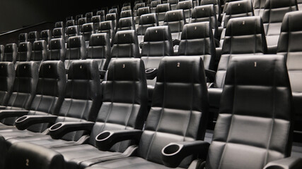 Rows of empty black leather cinema seats with cup holders and numbered backrests in a modern movie...