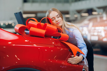 Happy young caucasian woman hugging his new first red car in showroom