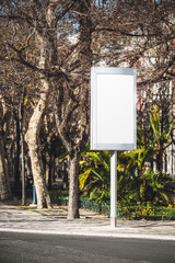A vertical telephoto of a blank billboard sign stands on a city sidewalk surrounded by bare trees and lush green foliage. The sign is ready for advertising in a public urban park or urban setting