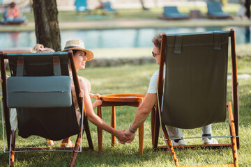 A couple holding hands while sitting on deck chairs near a pool, with a child in the background. The scene captures a moment of outdoor relaxation and family bonding