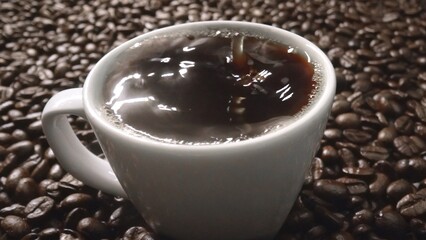 Close up of coffee bean drop in to hot espresso with pile of coffee seed. Macro of freshly roasted bean falling in to cup with aromatic smoke. Slow motion. Organic coffee seed concept. Comestible.