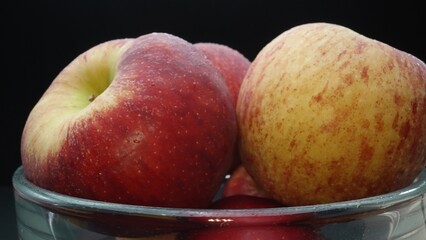 Close-up showcases a crisp bright red fresh apple nestled comfortably in a glass bowl. The black background creates a dramatic stage, highlighting the apple's flawless and smooth skin. Comestible.