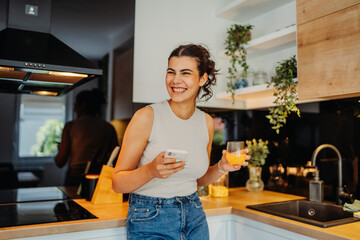 Young caucasian woman drinking orange juice and using phone in kitchen
