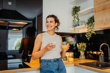 Young caucasian woman drinking orange juice and using phone in kitchen