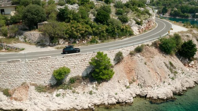 Auto with roof box drives along coastal road to lagoon with azure water. Highway between sea and rocky hillside creates fascinating views