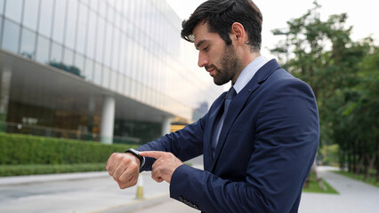 Skilled caucasian business man looking at time while standing at building. Attractive executive...