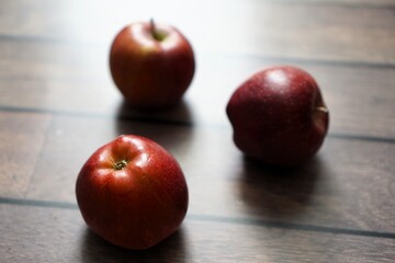 A simple arrangement of three red apples placed on a wooden floor, softly lit to highlight their natural sheen and color, creating a minimalist and organic composition.
