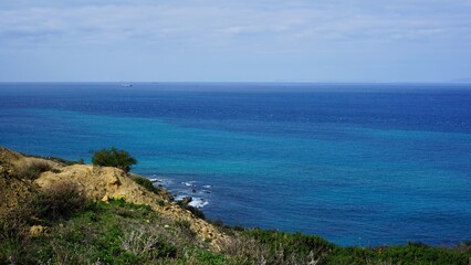 A scenic coastal view featuring a cliff covered in greenery, overlooking the turquoise waters of the ocean under a partly cloudy sky, Morocco.
