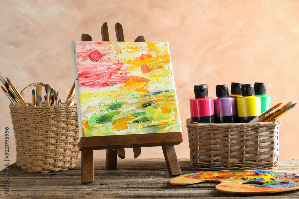 Poster Acrylic paints of different colors, small easel with abstract picture, palette and brushes on wooden table