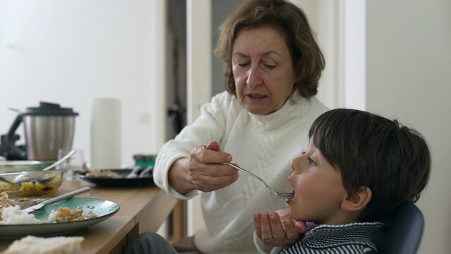 Grandmother feeding young boy a spoonful of food at the dining table, tender family moment, multi-generational care and love, homey atmosphere, child eating with grandparent assistance