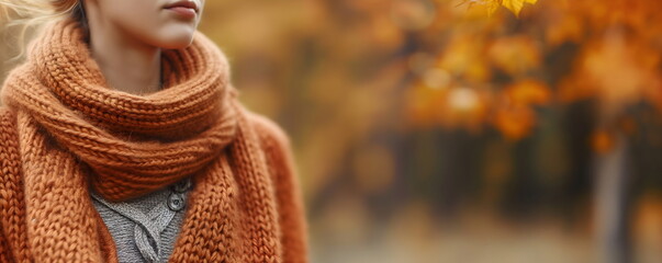 Close-up of woman's neck wrapped in orange knit scarf, autumn fashion detail with blurred fall background. Banner with copy space