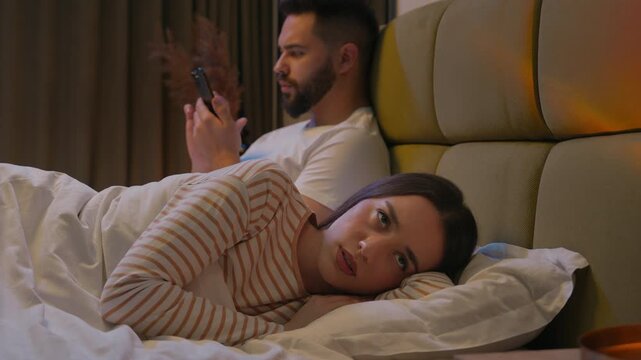Caucasian woman girlfriend lying in bed turned away man boyfriend glued mobile phone smartphone dissatisfied relationship attention addiction upset separate offended crisis female male stuck bedroom