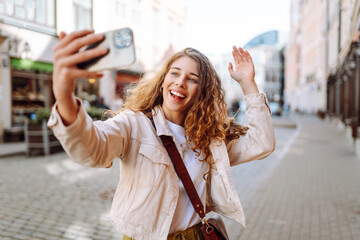 Optimistic young woman looking at camera with smile while taking selfie on city street. Technology, blogging, communication concept.