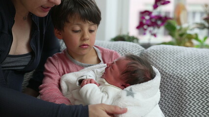 Mother and young boy sitting together, holding newborn sibling in white blanket, experiencing family bonding, sibling love, and parental care, cozy indoor setting, affectionate family moment
