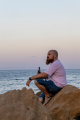 Bald man with a beard drinking a bottle of beer on the beach.