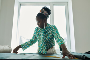 Young African American seamstress in green blouse measuring piece of textile while bending over...