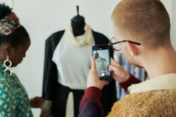 Rear view of male tailor with mobile phone taking photo or video of young African American seamstress and mannequin with attire