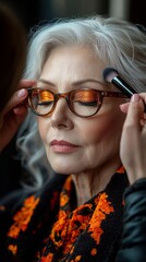 A 55-year-old woman with gray hair and glasses receives makeup application, showcasing vibrant orange eyeshadow in a studio.