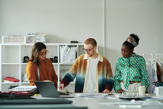 Three young intercultural fashion designers in smart casualwear standing by workplace and choosing textile for new attire