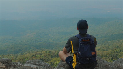 Man sitting on rock, enjoying panoramic view of lush green forest and distant horizon on foggy day, showcasing beauty of nature and thrill of adventure. Outdoor Adventure and Nature.