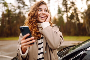 Young woman standing by the car and using phone. Car sharing, rental service or taxi app....