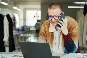 Young smiling man in casual apparel and eyeglasses speaking on mobile phone and using laptop while...