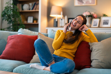 Obraz premium A cheerful Caucasian woman engages in a lively phone chat, comfortably seated on her living room sofa, adding warmth to the cozy domestic setting.