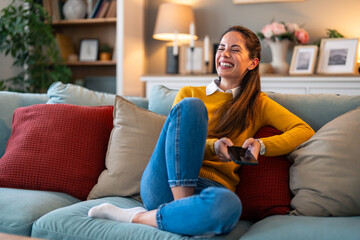 A cheerful Caucasian woman wearing a cozy yellow sweater smiles brightly as she interacts with her smartphone, comfortably seated on a couch in a homey living room setting.