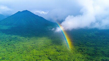   An aerial view of a rainbow in a lush green area, with a majestic mountain in the backdrop and fluffy clouds above