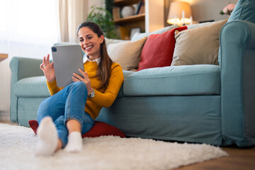 A cheerful woman enjoys a digital connection at home, engaging through video chat on her tablet while sipping coffee, seated comfortably by her couch.