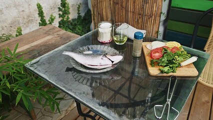 Outdoor preparation for fish barbecue with fresh vegetables and ingredients on a rustic glass table set for a summer grilling experience.