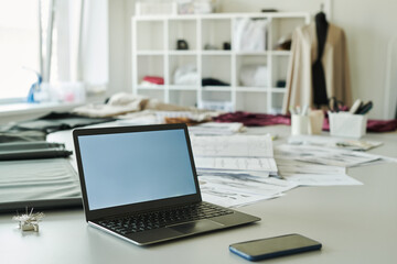 Laptop with blank screen standing in front of camera in the center of workplace of modern fashion designer or tailor in atelier