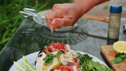 Person squeezing fresh lemon juice onto a seasoned fish barbecue with herbs and vegetables on a plate