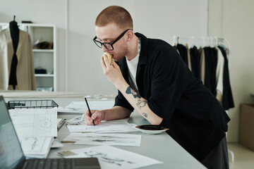 Young busy male fashion designer with pencil biting sandwich and drawing sketch of attire for new seasonal collection by workplace
