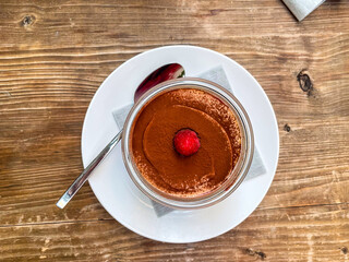 round glass jar with chocolate sprinkles and a raspberry on white cream on a wooden table, tiramisu, top down view