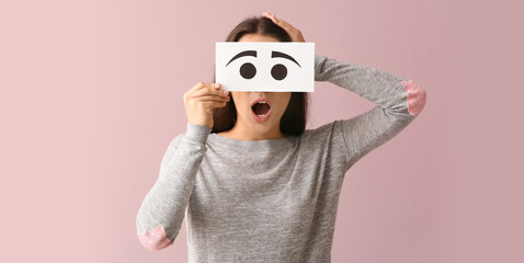 Emotional young woman hiding face behind sheet of paper with drawn eyes on color background