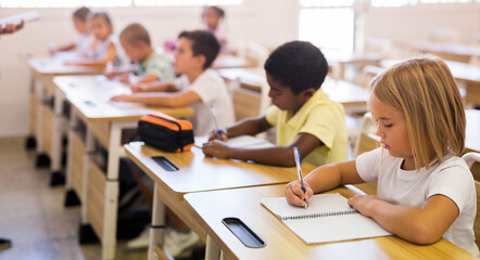 Diligent blonde preteen girl studying in classroom, listening to teacher and writing in notebook