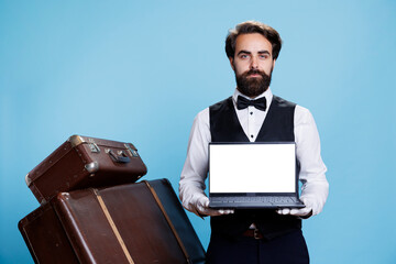 Bellboy presenting blank display on pc and advertising tourism industry, standing against blue background. Hotel concierge showing screen with white copyspace template on camera.