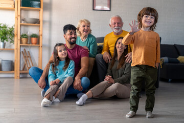 Diverse family with cheerful expressions sits together in a cozy living room; young boy stands in front, adding playful energy to the group portrait.