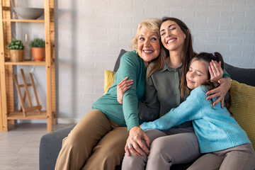 A cheerful elderly woman, her daughter, and young granddaughter share a joyous embrace on a cozy living room sofa, radiating happiness and familial love.