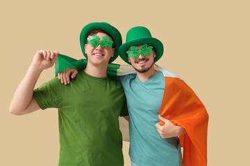 Happy young men in leprechaun's hats and party glasses with flag of Ireland hugging on beige background. St. Patrick's Day celebration