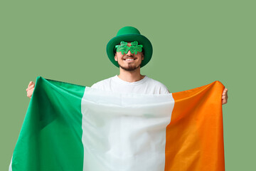 Happy young man in leprechaun's hat with party glasses and flag of Ireland on green background. St....