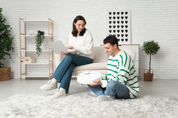 Young couple with modern robot vacuum cleaner and laptop in living room